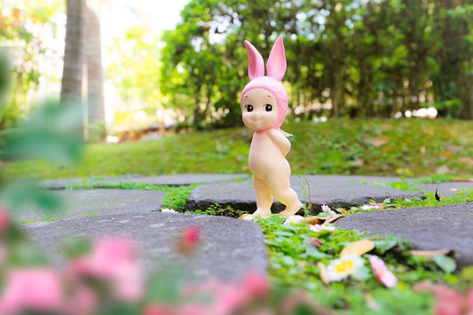 A Sonny Angel bunny standing on a pavement with his hands behind his back, holding a flower