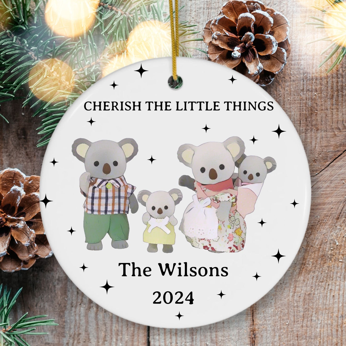Decorative Christmas ornament with cartoon koalas and text 'Cherish the Little Things' on a wooden background.