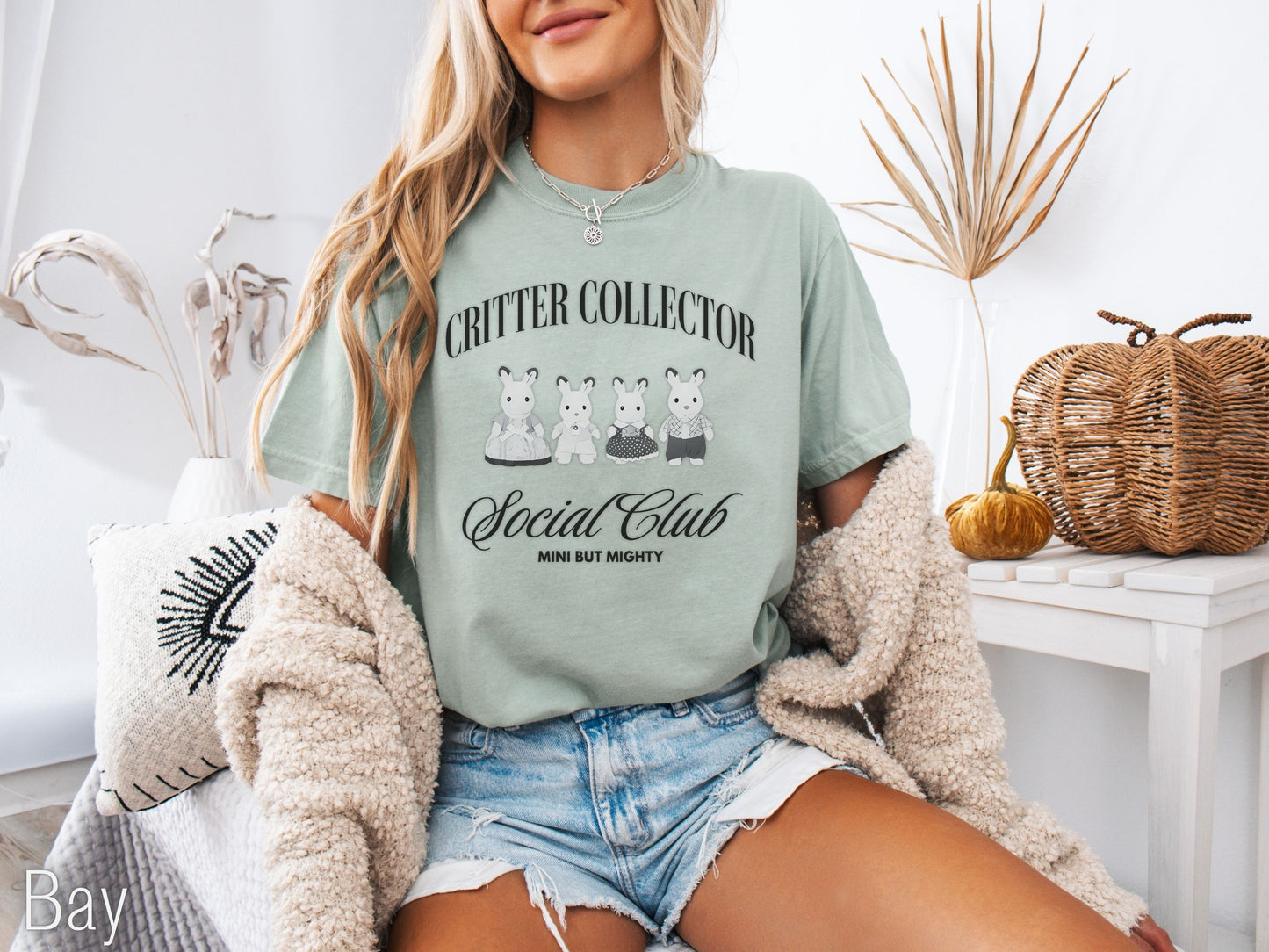 Woman wearing a bay t-shirt with 'Critter Collector Social Club' design, sitting in a cozy room.