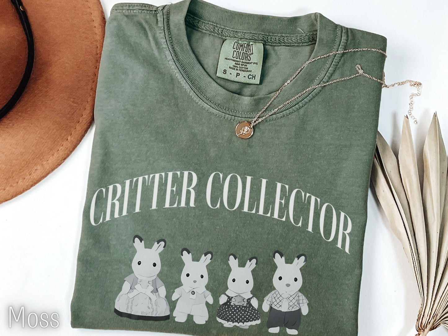Green t-shirt with 'Critter Collector' text and cartoon characters, placed on a white surface with a hat and dried leaves.
