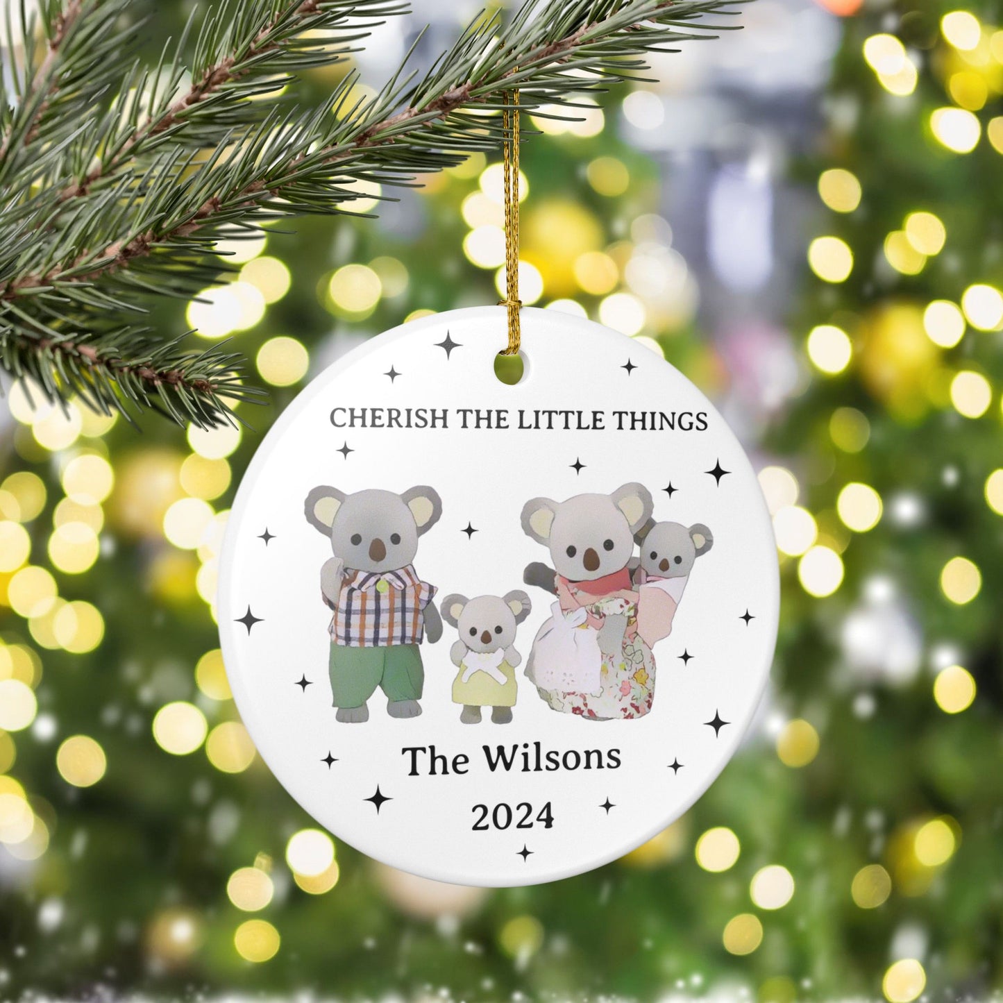 Decorative Christmas ornament with calico critter family of koalas and text 'Cherish the Little Things' on a blurred Christmas tree background.