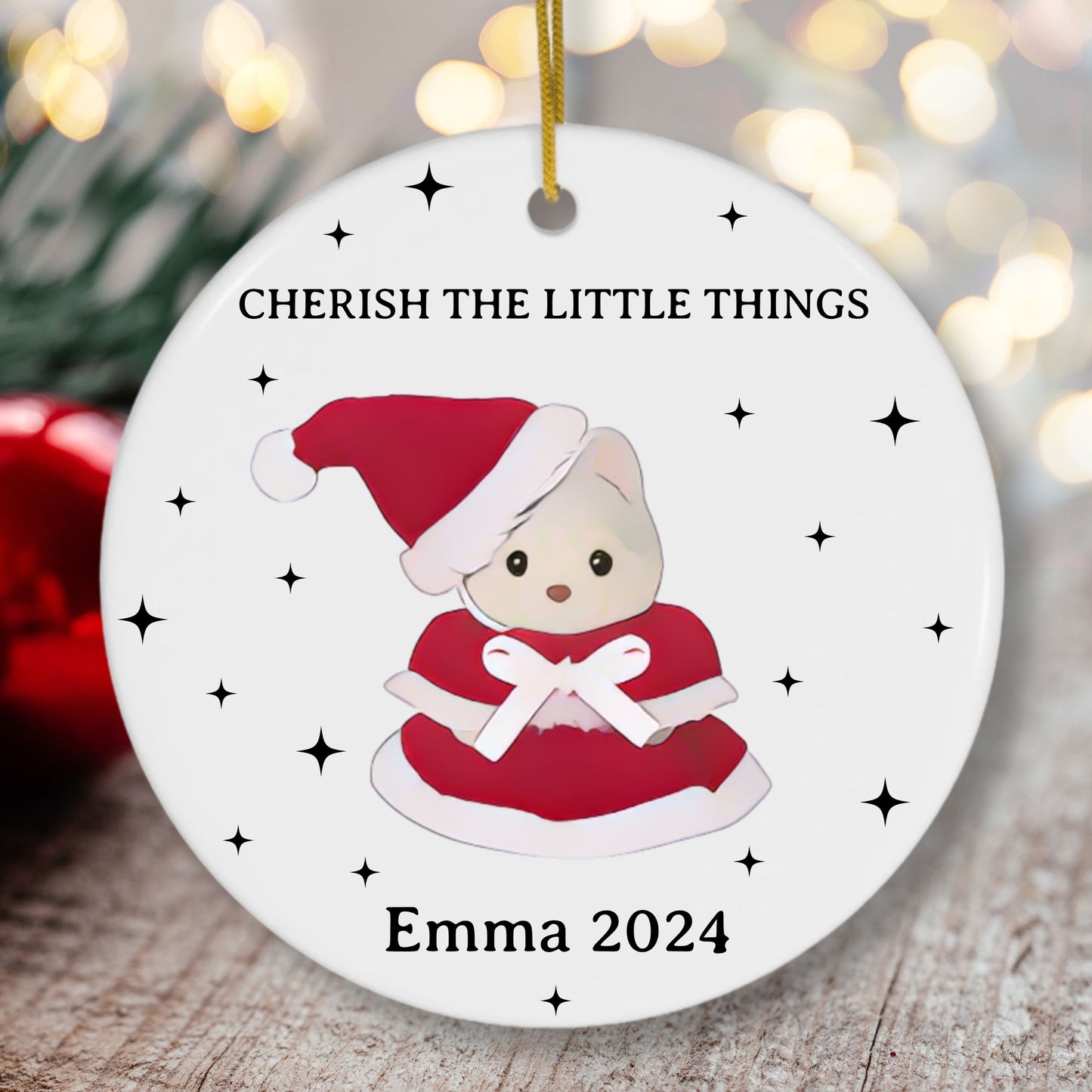 Christmas ornament with a calico critter bear character wearing a Santa hat and red dress, text 'CHERISH THE LITTLE THINGS' and 'Emma 2024', against a blurred festive background.