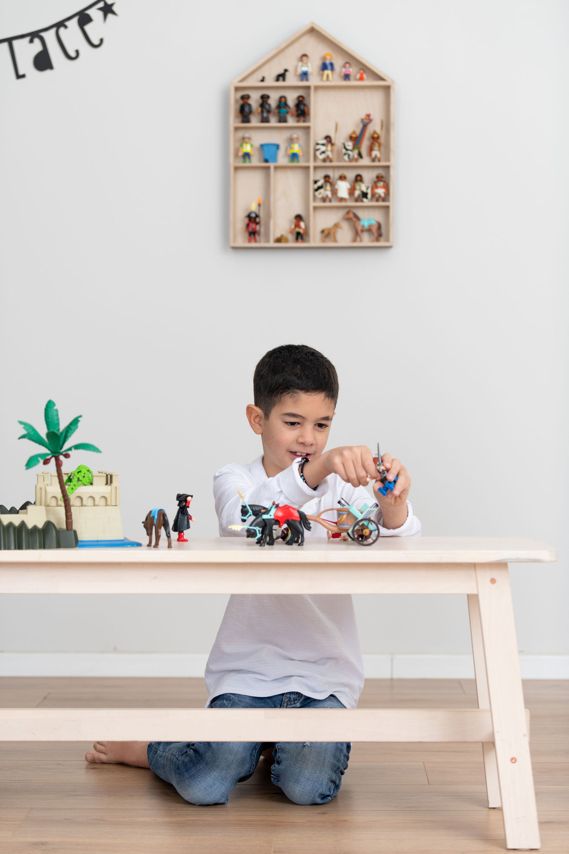 A sits near a bench and plays with Playmobil near a house-shaped toy display shelf that is hung on the wall