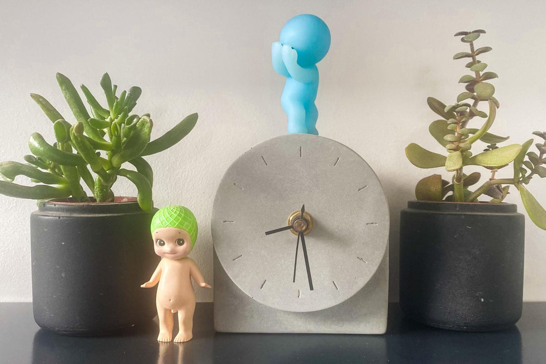 Smiski and Sonny Angel on a shelf with plants and a clock.