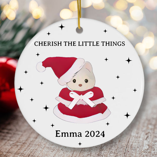 Christmas ornament with a calico critter bear character wearing a Santa hat and red dress, text 'CHERISH THE LITTLE THINGS' and 'Emma 2024', against a blurred festive background.