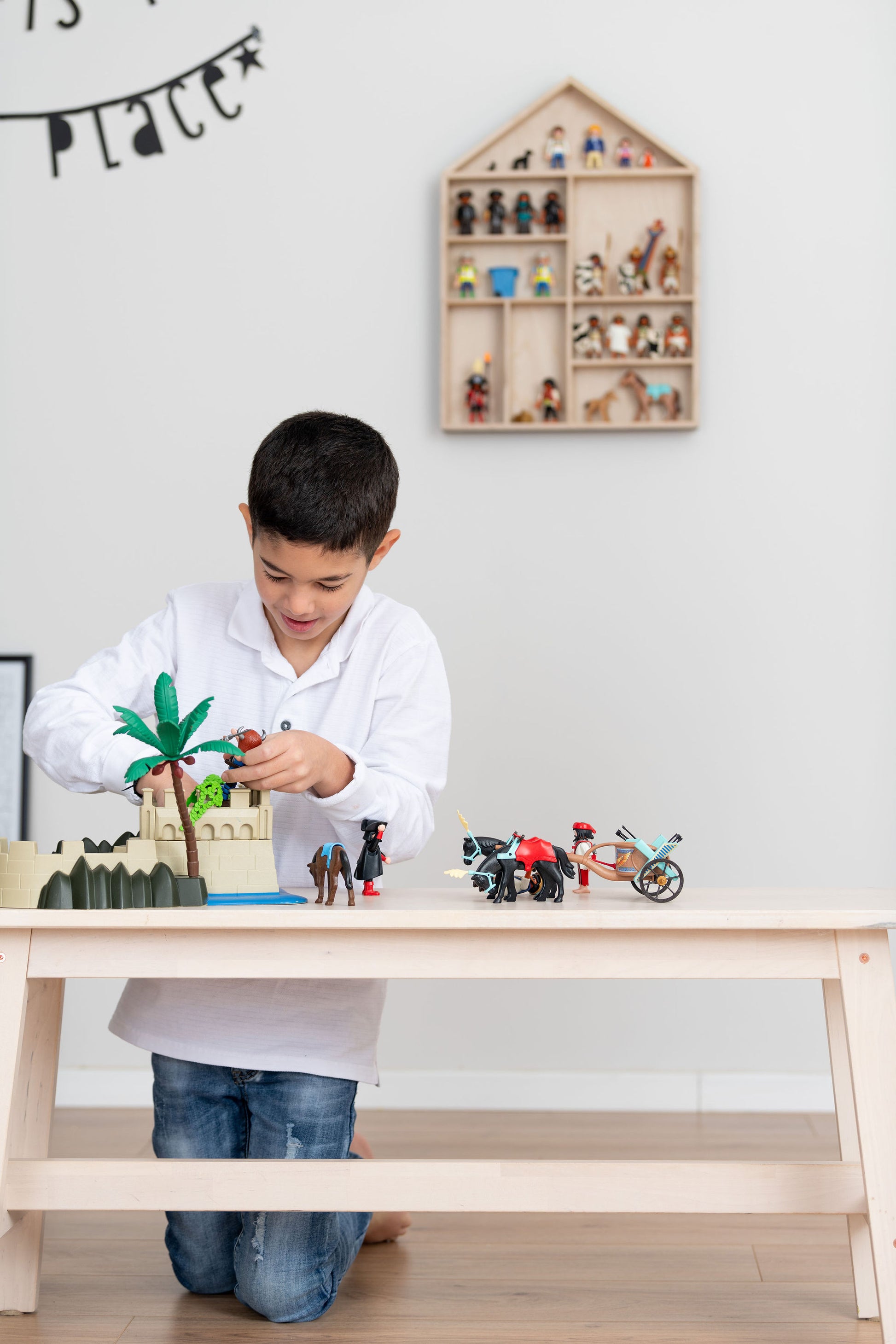 A boy sits near a bench and plays with Playmobil near a house-shaped toy display shelf that is hung on the wall