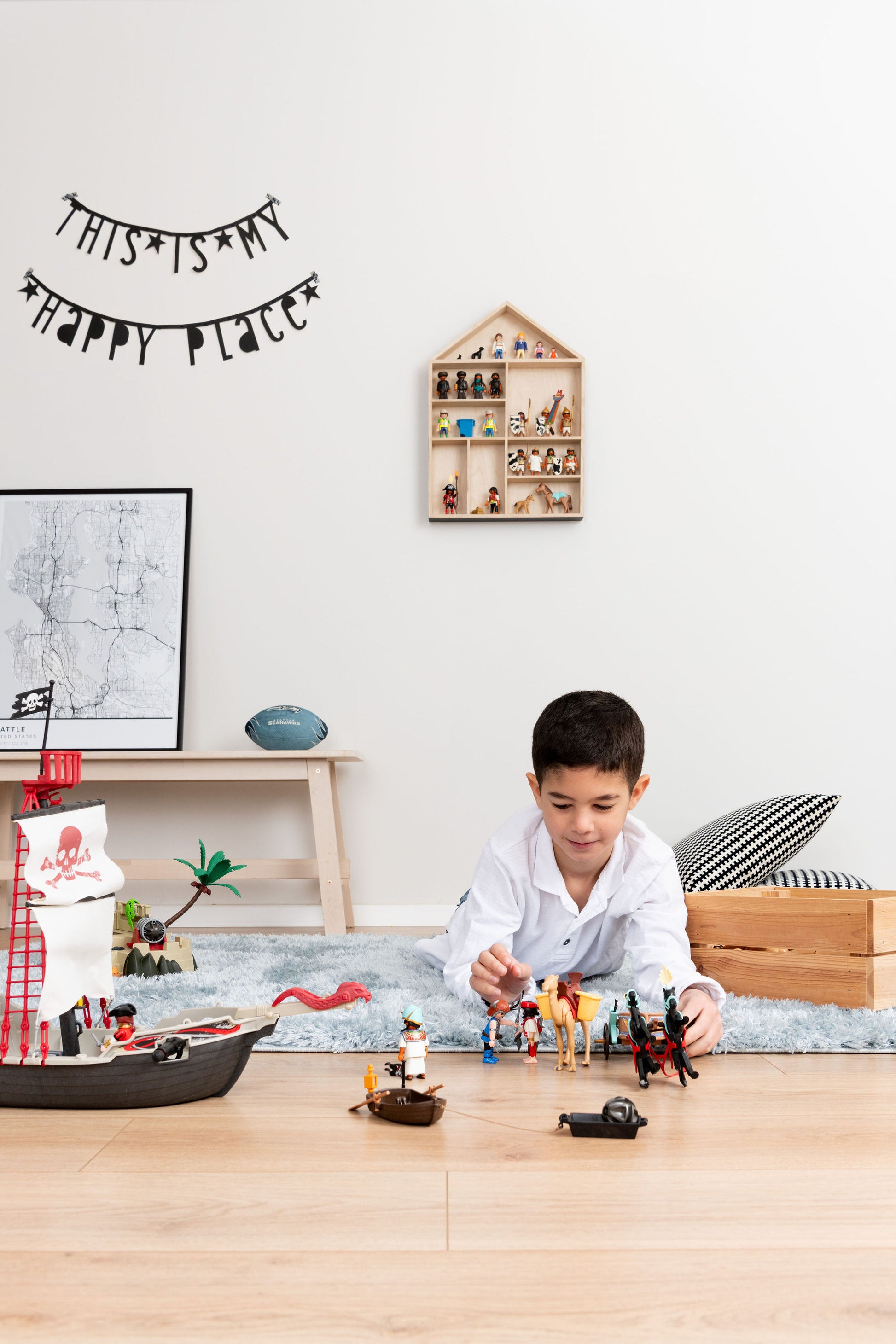 A boy laying on the rug is playing with Playmobil near a house shaped toy display shelf that is hung on the wall
